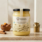 Chahal Agri Farms desi cow ghee glass jar on a stone counter with turmeric root, cinnamon and a brass milk pot